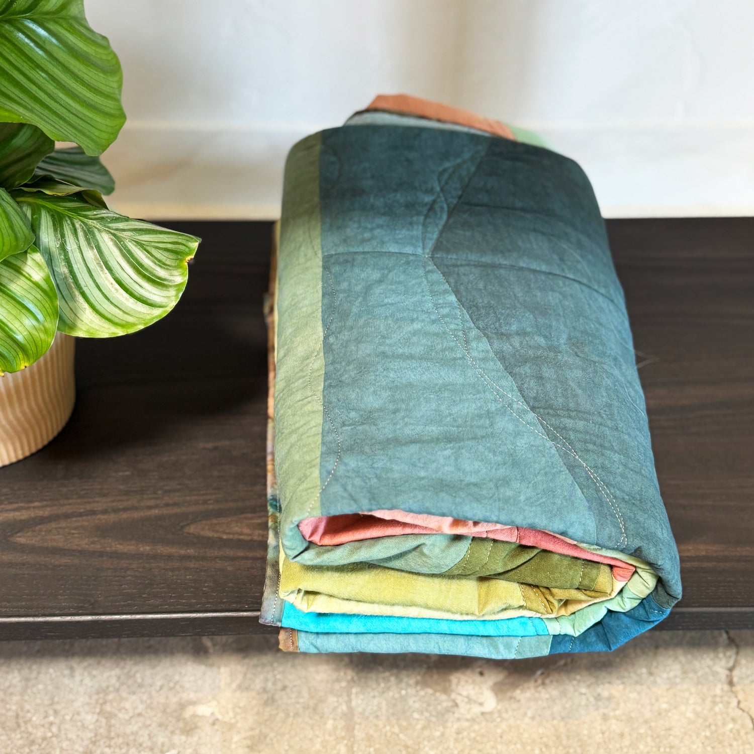 Folded quilted blanket on a wooden surface with a plant in the background